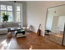 Wohnzimmer mit großen Fenstern, einem hellen Ecksofa, einem Sessel mit Metallrahmen und einem runden Hocker auf einem Teppich. Im Raum befinden sich eine Zimmerpflanze, eine an die Wand gelehnte Gitarre und ein großer Spiegel, der das Zimmer reflektiert. Der Boden ist aus Parkett, und im Hintergrund ist ein angrenzender Raum sichtbar.