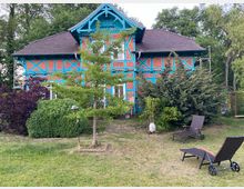 Ein zweistöckiges Fachwerkhaus mit roten Ziegeln und türkisfarbenen Holzbalken, umgeben von Bäumen, Sträuchern und einem gepflegten Garten. Im Vordergrund stehen zwei Liegestühle auf einer Rasenfläche.