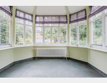 Heller, leerer Erkerraum mit großen, umlaufenden Sprossenfenstern und halbtransparenten Raffrollos. Die Wände sind hellgrün, am Fenster steht ein weißer Heizkörper, und der Boden ist mit dunkelgrauen Fliesen ausgelegt; draußen sind Bäume und Nachbarhäuser zu sehen.