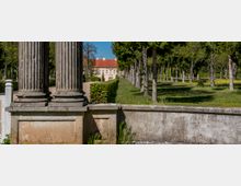 View from a stone terrace with large fluted classical columns, looking along a manicured garden with clipped hedges and rows of trees. A pale historic manor with a red-tiled roof stands in the distance beyond the formal grounds.