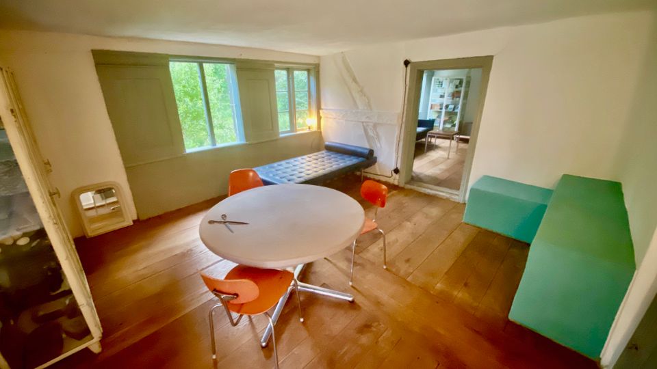 Heller Wohnraum mit breiten Holzdielen, weiß verputzten Wänden und großen Fenstern mit grünen Klappläden. In der Mitte stehen ein runder Tisch mit orangefarbenen Stühlen, dazu eine Liege an der Fensterwand und türkisfarbene Sitzblöcke neben einer offenen Tür zu einem Nebenraum.