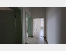 Schmaler, leerer Innenflur mit weißen Wänden, hellgrünen Holztüren und einem einfachen Bodenbelag. Am Ende öffnet sich der Gang zu einem hellen Raum mit großem Rundbogenfenster und einem Heizkörper darunter.