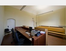 Ein schlicht eingerichteter Gerichtssaal mit einem großen Holztresen im Vordergrund, ausgestattet mit Computer, Mikrofon und Drucker. Im Hintergrund befinden sich Sitzbänke aus Holz, helle Wände, eine Uhr und weiße Türen.