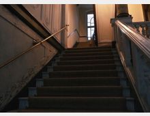 Treppenaufgang in einem historischen Gebäude mit dunklem, mit Teppich ausgelegtem Treppenbelag, verzierten Wänden und Geländern aus hellem Stein. Im Hintergrund ist ein großes Fenster mit Rundbogen erkennbar.