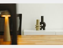 Minimalistischer Innenraum mit einem weißen Schrank, auf dem zwei moderne Vasen in geometrischen Formen stehen. Im Vordergrund sind ein Holztisch und ein Stuhl mit angerautem Netzdesign zu sehen, links eine sanft leuchtende Lampe.