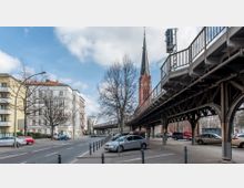 Straßenszene in einer Stadt mit Wohngebäuden links und einer Hochbahntrasse rechts. Im Hintergrund ragt ein roter Kirchturm mit Spitzdach und Uhr in den Himmel, während Autos auf der Straße und unter der Hochbahn geparkt sind.