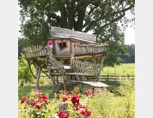 Ein rustikales Baumhaus aus Holz mit schrägem Dach und Geländern, das auf Stelzen steht; im Vordergrund befinden sich blühende rote Rosen, während im Hintergrund eine weitläufige grüne Wiese und ein Zaun zu sehen sind. Ein Kind steht auf der Holztreppe, die zum Baumhaus führt.