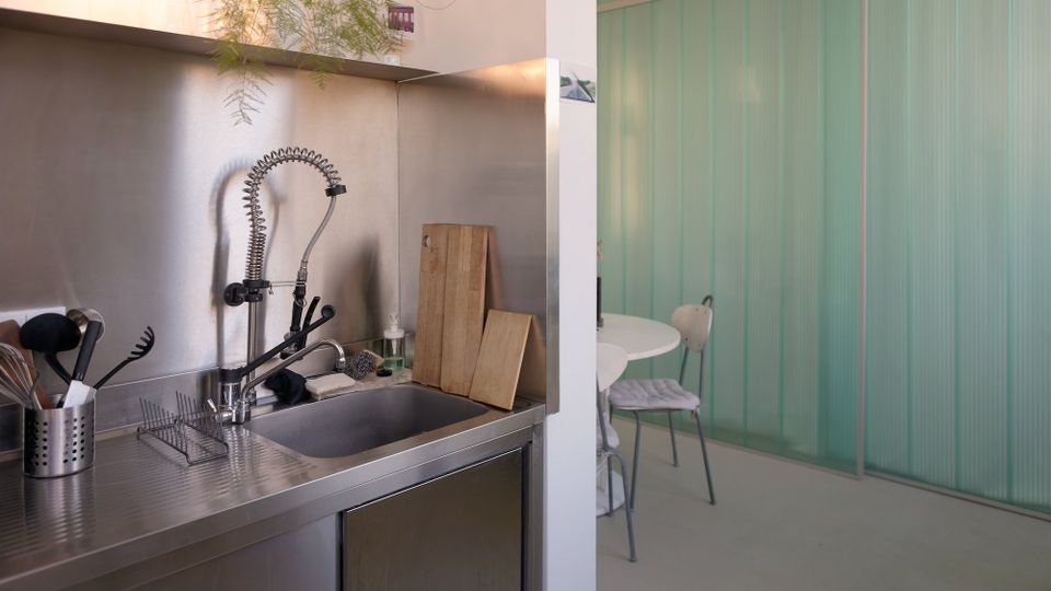 Moderne Küchenzeile aus Edelstahl mit tiefem Spülbecken, hoher Spiralfeder-Armatur, Abtropfgitter und Küchenutensilien vor einer Metallrückwand. Rechts öffnet sich der Raum zu einem Essbereich mit rundem Tisch vor einer mintgrünen, gerippten Trennwand.