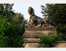 Steinfigur einer liegenden Sphinx auf einem dekorativen Sockel in einem grünen Garten, umgeben von Bäumen und Sträuchern. Die Statue zeigt verwitterte Details und ist von natürlichem Licht beleuchtet.