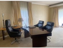 Elegantes Büro mit einem dunklen Holztisch, drei schwarzen Ledersesseln und einem kleinen Beistelltisch mit Lampe. Der Raum hat cremefarbene Wände, helle Vorhänge und große Fenster, die viel Tageslicht einlassen.