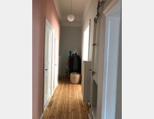 Ein schmaler Flur in einer Wohnung mit hellem Holzboden und Wänden, die teils rosa und teils weiß gestrichen sind. Am Ende des Flurs steht eine Garderobe mit aufgehängten Kleidungsstücken, daneben ein geflochtener Korb; an der Decke hängt eine runde Lampe.