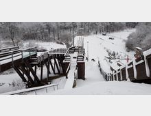 Eine verschneite Skisprungschanze mit Holzkonstruktionen, Treppen und Geländern, eingebettet in eine winterliche Waldlandschaft. Im Hintergrund sind ein schneebedecktes Stadion und umliegende Anlagen zu sehen.