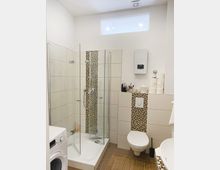 Helles Badezimmer mit Glasdusche, weiß gefliesten Wänden und Mosaikfliesen-Akzenten. Im Raum befinden sich ein Wand-WC, eine Waschmaschine, ein kleines Fenster oben an der Wand sowie eine Ablage mit Toilettenartikeln.