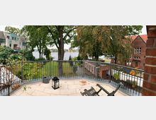 Blick von einer erhöhten Terrasse mit schmiedeeisernem Geländer auf einen Garten mit Bäumen und Rasen, dahinter ein See mit Steg. Auf der rechten Seite befindet sich ein rotes Backsteingebäude, links ein Wohnhaus mit Balkonen.