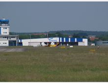 Flugplatz mit einem kleinen Hangar in blau-weißer Farbe, einem Kontrollturm links und einer gelben Propellermaschine im Vordergrund. Im Hintergrund sind Wälder und vereinzelte Gebäude sichtbar.