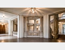 Großzügiger, klassisch gestalteter Innenraum mit hohen Steinpfeilern, Balustraden und Rundbögen. Der Raum hat verzierte Holzdetails, eine elegante Treppe und indirekt beleuchtete Lampen an der Decke.