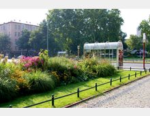 Begrünte Stadtfläche mit Blumenbeeten und einer überdachten Bushaltestelle aus Glas. Im Hintergrund ein mehrstöckiges Wohngebäude, Bäume sowie eine Straße mit Verkehrsschildern und Gehwegen.