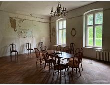 Ein einfacher, leerer Raum mit abgenutzten, teils abgeblätterten Wänden, großen Fenstern, die Tageslicht hereinlassen, und einem Holztisch, der von mehreren Stühlen umgeben ist. An der Decke hängt ein Kronleuchter, und der Holzfußboden ist leicht abgenutzt.