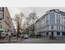 Straßenszene in einer Wohngegend mit mehrstöckigen Altbauten; links ein gelbliches Gebäude mit Geschäften und einem Straßenmarkt, rechts ein weiß-blaues Eckgebäude mit Ladengeschäften im Erdgeschoss. Im Hintergrund nackte Bäume und geparkte Autos entlang der Straße.