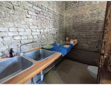 Eine einfache Küche mit unverputzten Backsteinwänden, zwei Edelstahlspülbecken und einer Holzarbeitsplatte. Auf der Arbeitsplatte befinden sich Geschirr, Gläser, eine blaue Tischdecke und Küchenutensilien.