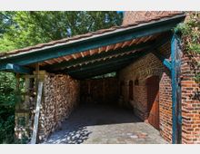 Überdachter Holzlagerraum mit Backsteinwänden, einem Ziegeldach auf blauen Holzbalken und gestapeltem Brennholz an der Wand. Im Hintergrund sind Bäume und im Vordergrund eine gepflasterte Fläche sichtbar.