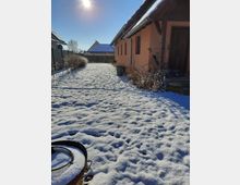 Schneebedeckter Garten mit einem pfirsichfarbenen Haus auf der rechten Seite, einer überdachten Eingangstür und kahlen Sträuchern entlang der Fassade. Im Vordergrund ist eine Feuerschale sichtbar, im Hintergrund weitere Häuser und ein blauer Himmel mit Sonne.