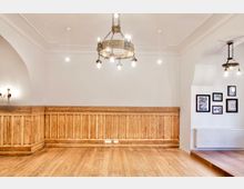 Heller Innenraum mit weißer Decke und Wänden, umlaufender Holzvertäfelung und breitem Holzdielenboden. Von der Decke hängen historische Kronleuchter; rechts sind gerahmte Bilder über einem Heizkörper neben einer kleinen Stufe zu sehen.