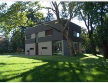 Modernes zweistöckiges Wohnhaus mit klaren Linien und grauer Fassade, umgeben von einer gepflegten Rasenfläche und hohen Bäumen in einer parkähnlichen Umgebung. Große Fenster prägen die Architektur, während Sonnenlicht durch die Bäume auf das Grundstück fällt.