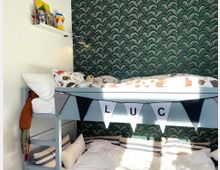 Ein Kinderzimmer mit einem Hochbett aus hellblauem Holz. Die Bettwäsche hat ein Tiermotiv, und an der Bettrahmenkante hängt eine Wimpelkette mit Buchstaben. Im Hintergrund ist eine grüne Tapete mit fächerartigen Mustern zu sehen, und an der weißen Wand hängt ein Regal mit Spielsachen und Büchern.