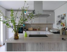 Moderne Küche mit heller Holzarbeitsplatte und weißen Schränken. Im Vordergrund steht ein großer Zweig mit bunten Ostereiern in einer Vase auf der Kücheninsel, im Hintergrund sind ein eingebauter Backofen, Küchengeräte und Wandregale zu sehen.