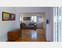 Wohnraum mit heller Einrichtung, einem Parkettboden und weißen Wänden. An den Wänden hängen farbige Bilder, links steht eine antike Holztruhe, im Hintergrund eine graue Eckcouch vor einem Fenster.