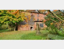 Backsteinbau mit Holzelementen, umgeben von Bäumen und einer Wiese. Im Vordergrund eine kleine Steinfigur und eine grüne Sitzbank.