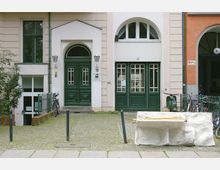 Fassade eines mehrstöckigen Gebäudes mit beigen Wänden und dunkelgrünen Holztüren. Links und rechts des Eingangsbereichs stehen Laternen, davor befinden sich Fahrräder und eine weiße Skulptur auf einem gepflasterten Vorplatz.