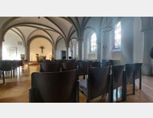 Innenansicht einer kleinen Kirche mit Gewölbedecken und großen Bögen. Im Vordergrund befinden sich leere Stuhlreihen, im Hintergrund ein schlichter Altar mit Holzkreuz sowie Schmückungen mit Blumen; an der Seite sind bunte Kirchenfenster sichtbar.
