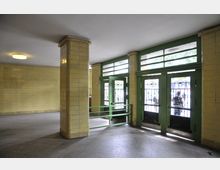 Eingangsbereich mit gelb gefliesten Wänden, grünen Metalltüren mit Glaseinsätzen und dekorativen Gittern. Der Boden ist mit grauen Fliesen bedeckt, und durch die Türen ist eine Außenszene mit Personen erkennbar.