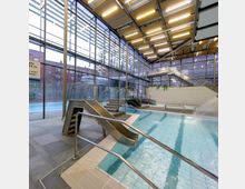 Innenansicht eines modernen Hallenbads mit großen Glaswänden, die den Blick nach draußen ermöglichen. Sichtbar sind Wasserrutschen, ein Wasserfall im Becken sowie Sitzgelegenheiten, alles umgeben von metallischen und gefliesten Oberflächen.