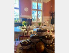 Gemütlicher Innenraum eines Cafés mit hohen Fenstern, durch die Tageslicht hereinfällt, und pastellfarben gestrichenen Wänden. Im Vordergrund ein Teller mit Schokoladenmuffins, im Hintergrund ein runder Tisch mit Blumen und Dekorationen.