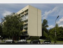 Mehrstöckiges Wohngebäude mit modernem, schlichtem Design und leicht geneigter Fassade, umgeben von Bäumen. Im Vordergrund parken mehrere Autos entlang einer Straße, eine Laterne ist sichtbar.