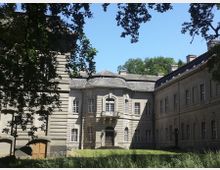 Historisches Gebäude mit klassizistischer Architektur, bestehend aus hellen Steinmauern und symmetrischen Fenstern, umgeben von Bäumen und einer Rasenfläche. Ein mittlerer Flügel mit einem kleinen Balkon und einer Eingangsportaltreppe steht im Zentrum des Gebäudekomplexes.
