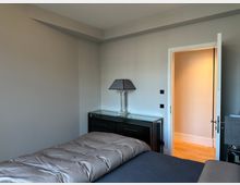 Schlichtes Schlafzimmer mit grauen Wänden und Bett im Vordergrund; an der Wand steht eine schwarze Kommode mit Glasplatte und Tischlampe. Rechts führt eine offene Tür in einen hell beleuchteten Flur mit Holzfußboden.