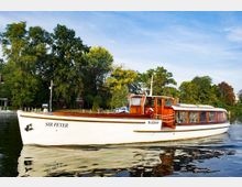 Weißer Ausflugsdampfer mit dem Schriftzug „Sir Peter“ und Holzapplikationen, der auf einem ruhigen Fluss vor einer Uferlandschaft mit grünen Bäumen und Häusern fährt. Der Himmel ist klar und blau, und das Wasser spiegelt das Boot.