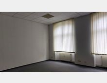 Leerer Raum mit weiß gestrichenen Wänden, Teppichboden und einer abgehängten Decke mit Rasterleuchten. Zwei große Fenster mit vertikalen Lamellenjalousien lassen Tageslicht herein, darunter befinden sich Heizkörper.