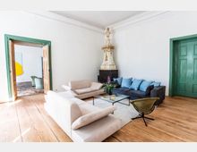 Helles Wohnzimmer mit Holzdielenboden, einer modernen Sitzgruppe aus beigen und grauen Sofas, einem Glastisch und einer dekorativen, historischen Kaminverkleidung in Weiß und Gold. Im Hintergrund offene Doppeltüren mit grünen Türrahmen führen in einen weiteren Raum.