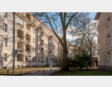 Mehrstöckiges Wohngebäude aus der frühen Moderne mit abgerundeten Balkonen und symmetrischen Fassadendetails, umgeben von kahlen Bäumen und einem kleinen gepflasterten Weg. Im Vordergrund befindet sich ein Schild für eine verkehrsberuhigte Zone.