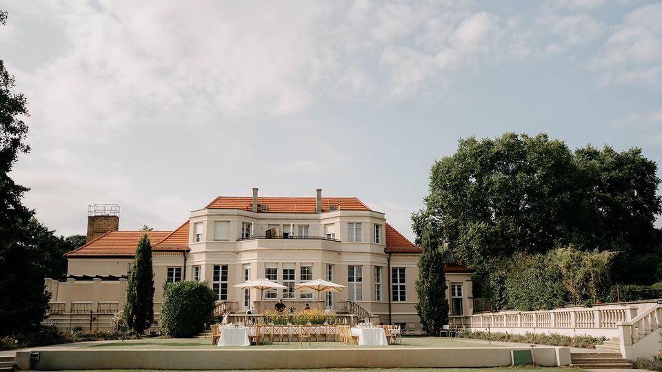 Große, helle Villa mit rotem Ziegeldach und symmetrischer Fassade, davor eine breite Terrasse mit gedeckten Tischen und Sonnenschirmen. Im Vordergrund liegt eine weitläufige Rasenfläche, seitlich eingefasst von Bäumen, Hecken und einer Balustrade.