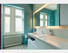 Modernes Badezimmer mit türkisfarbenen Mosaikfliesen an den Wänden und einem wandbreiten beleuchteten Spiegel. Im Vordergrund befindet sich ein Waschbecken mit einer glänzenden Armatur, ein Heizkörper unter einem großen Fenster und ein mit Fliesen belegter Waschtisch.