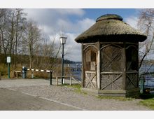 Ein hölzerner Pavillon mit strohgedecktem Dach steht an einem gepflasterten Weg nahe einem See. Im Hintergrund sind Bäume ohne Blätter, eine Schranke und ein Steg, der ins Wasser führt, sichtbar.