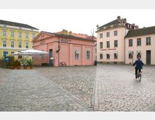 Gepflasterter Platz mit historischen Gebäuden, darunter ein gelbes und mehrere rosa Häuser, eines mit der Aufschrift „Gaststätte zur Ratswaage“. Im Vordergrund mehrere Blumentöpfe, rechts fährt eine Person mit einem Fahrrad.