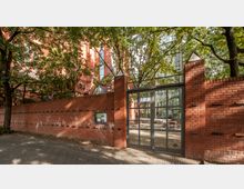 Rote Backsteinmauer mit einem Eisentor, dahinter ein Hof mit Bäumen und einem modernen Glasgebäude. Im Hintergrund ist ein historisches Backsteingebäude mit gotischen Fenstern sichtbar.