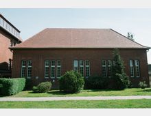 Backstein-Gebäude mit Satteldach und mehreren hohen, schmalen Fenstern in regelmäßiger Anordnung. Davor befindet sich eine Rasenfläche mit Sträuchern und einem Weg aus hellem Pflaster.